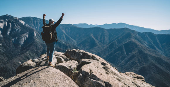 Hombre de espalda con los brazos alzados sobre la cima de una roca mirando una cadena montañosa
