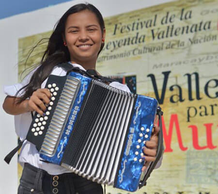 niña sonriente, de cabello largo, de unos 13 años, interpretando el acordeón delante de la propaganda del festival