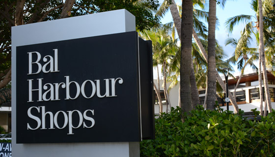Se ve el letrero negro con letras blancas que dice ¨Bal Harbour Shops¨, en el fondo plameras, árboles y el techo de una casa  