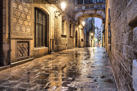 Anocheciendo, se ve un callejón del Barrio Gótico, con pequeños farole en las paredes alumbrando
