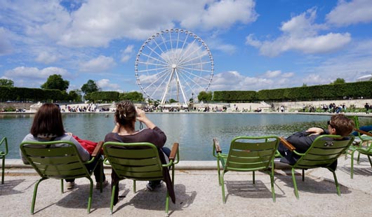 En primer plano personas sentadas en sillas verdes, frente a un lago. A lo lejos, se ve una rueda panorámica y pinos alrededor del parque. 