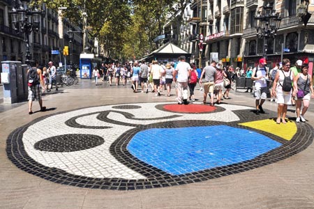 Se ve una avenida peatonal en medio de edificios, personas caminando y una figura en el suelo a base de cerámica de colores por la que las personas pueden caminar