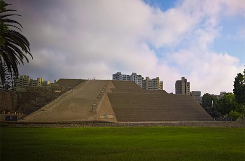 
Se ve un gran campo de césped y en el fondo una pirámide de tres niveles. Detrás, se ven diferentes edificios de la ciudad