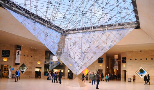 Salon en el que se encuentra una piramide invertida en vidrio y metal, que se encuentra en la punta con otra piramide pequeña en el piso.