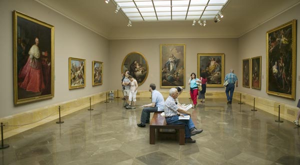 Se ve la sala de un museo, con diferentes pinturas con marcos dorados en las paredes, una silla de madera en medio en donde están sentadas varias personas, otras caminando ven las obras.