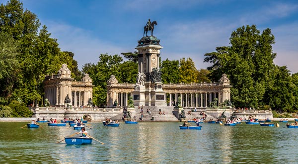 De día, se ve un lago rodeado de árboles en el que hay personas en canoas, al fondo se ve el monumento de Alfonso