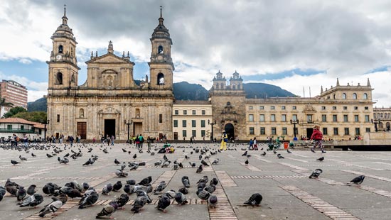 3.De día, se ve una plazoleta llena de palomas recogiendo maíz del piso y personas caminando entre ellas, al fondo, una catedral y construcciones coloniales. 