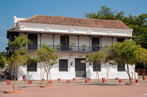 Vista de la casa de la Aduana, una arquitectura antigua, techo en ladrillo, fachada color blanco, rodeada de árboles 