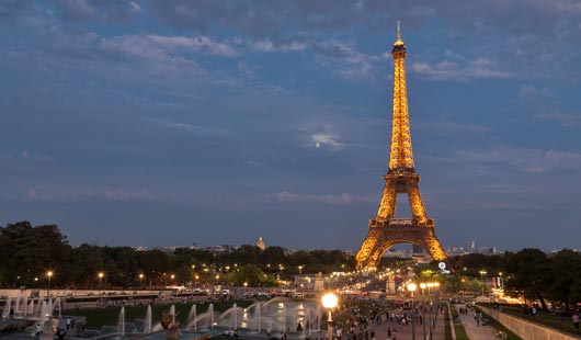 Vista nocturna de la torre Eiffel desde una calle peatonal con fuentes y grama, llena de gente