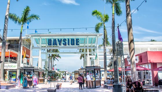 De día, se ve un arco con el nombre ¨bayside¨que da la entrada al mercado, donde se ven diferentes tiendas, casetas, sillas y personas caminando 