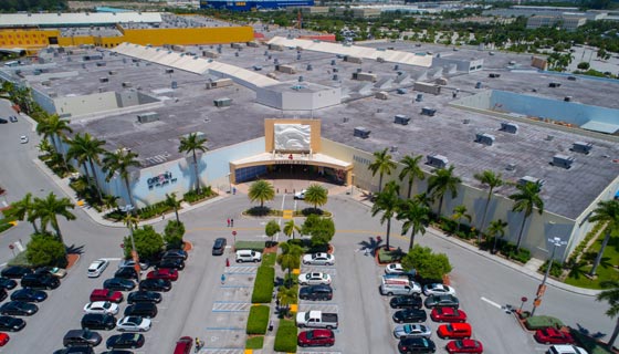 Vista aérea del exterior de un centro comercial rodeado de palmeras. En frente su parqueadero lleno de carros