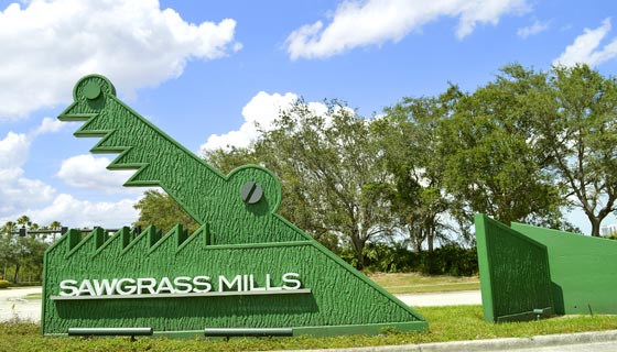 Se ve el aviso de centro comercial: en hierro una figura lineal de la cabeza de un cocodrilo con la boca abierta, y el nombre ¨sawgrass mills¨en ella. Al fondo pasto y árboles